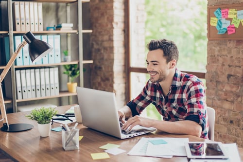 Cheerful young brunet freelancer is smiling, typing on his laptop in nice modern work station at home